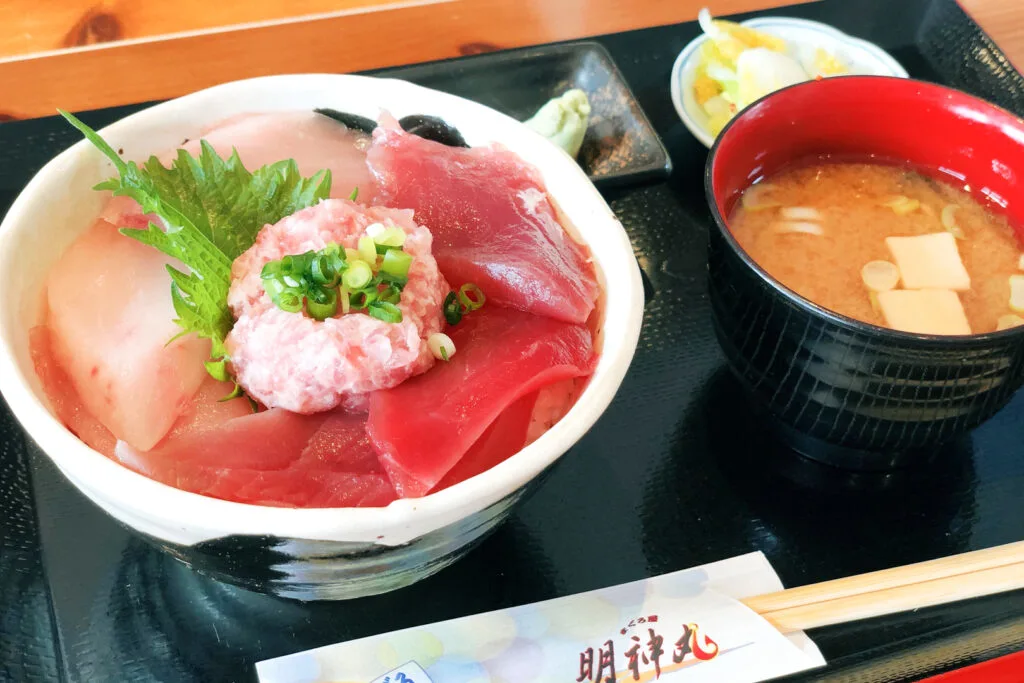 A maguro-don rice bowl topped with various cuts and types of tuna on a tray with wasabi, pickles, a bowl of miso soup and a pair of disposable chopsticks in a paper sleeve at the front.