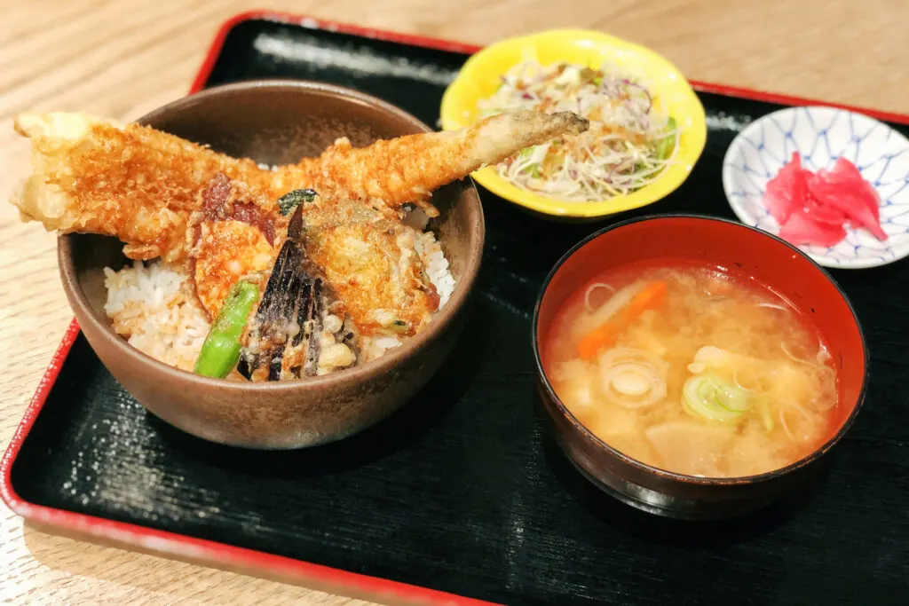 A half-sized anago tendon set, which includes a bowl of rice topped with deep-fried anago conger eel and tempura vegetables, a bowl of miso soup, a small side salad and some pickles. They are all sitting on a black lacquered wooden tray with a red rim, which is sitting on a light natural-colored wooden table.