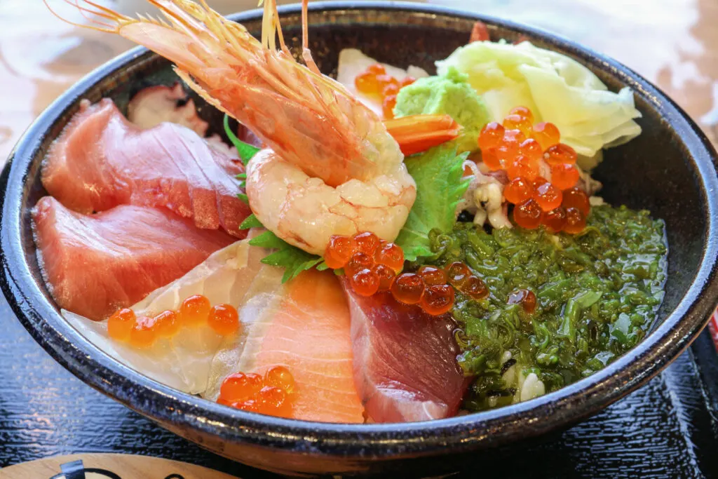 Okasei's Premium Onagawa don in a black wooden lacquered bowl on a matching black tray. The dish is topped with so much seafood that you can't see the rice under it. In the center is an elegantly twisted prawn atop a perilla leaf.