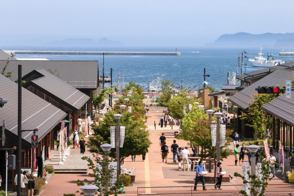 The SEAPAL-PIER area of Onagawa's Michi no Eki. There are tenant shops on either side with a tree-lined paved pedestrian walkway down the center leading to the ocean.