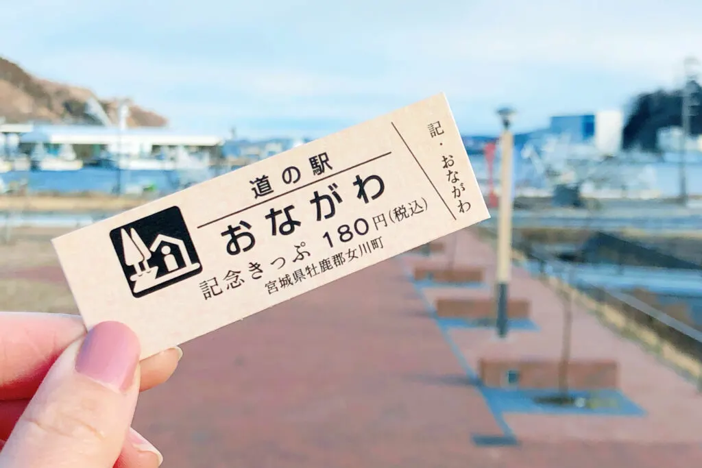 A left hand with pink nail polish holds out an Onagawa Michi-no-eki Commemorative Ticket with the port in the background.