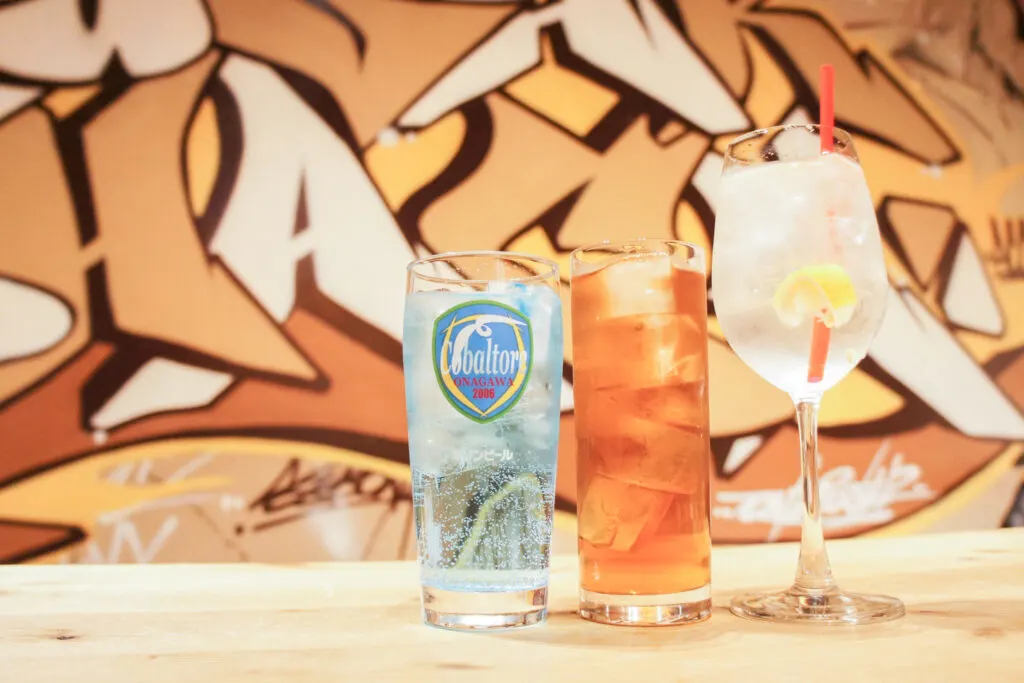 A Cobaltore Highball, a Reggae Punch and a Sake Squash lined up on a table with orange-colored graffiti art on the wall behind it.