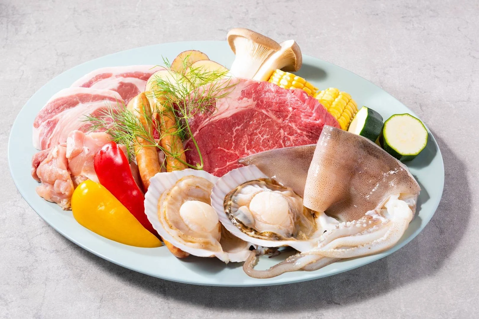 A tebura BBQ plate ready to be grilled. The plate has scallops. squid, various cuts of meat, peppers, corn, mushroom, zucchini and sweet potato, and is garnished with a sprig of fennel.