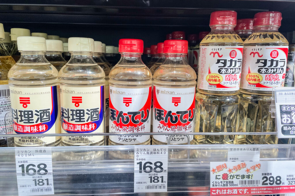 Mirin on a shelf at a supermarket in Tokyo. There are three products in view with two rows of bottles for each one. From left to right, we have Jozo chomiryo (181 yen with tax), Mirin-fu chomiryo for the same price, and Hon Mirin at the end for 327 yen.