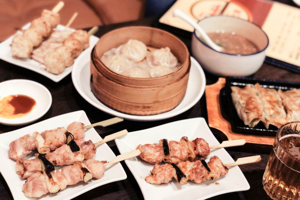 A full table of dishes, including various plates of chicken skewers and some steamed dumplings in a bamboo steamer basket in the center.