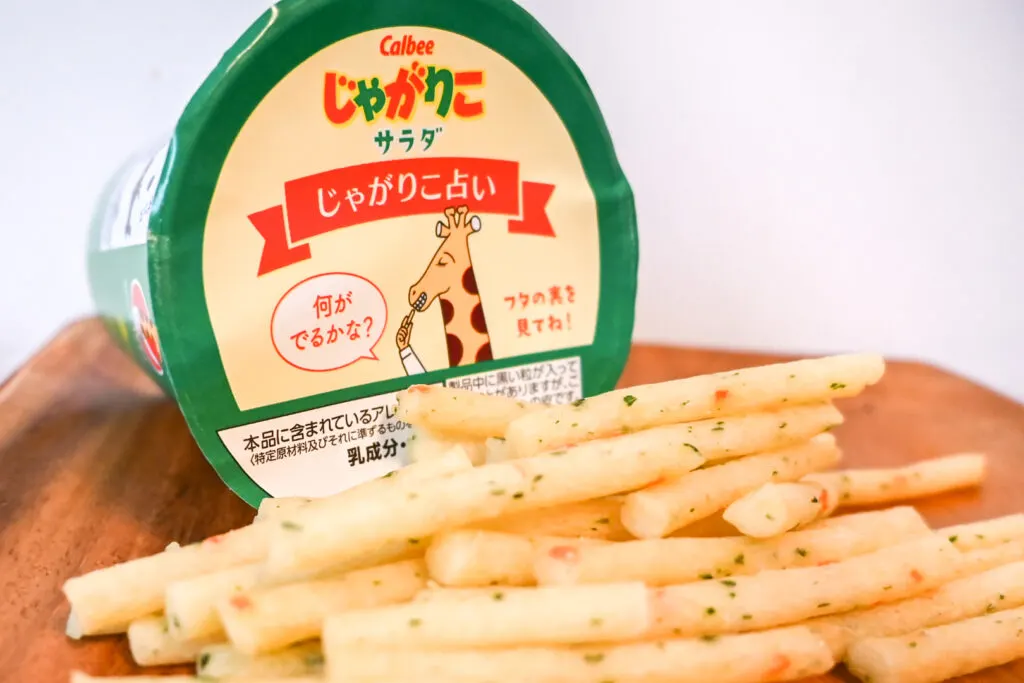 Jagarico potato sticks come in a cup similar to instant ramen. It has the same kind of peel off lid as well. In the image, the cup has been placed on its side on a wooden tray to show the lid and in front of it are the potato sticks with little bits of green and orange 