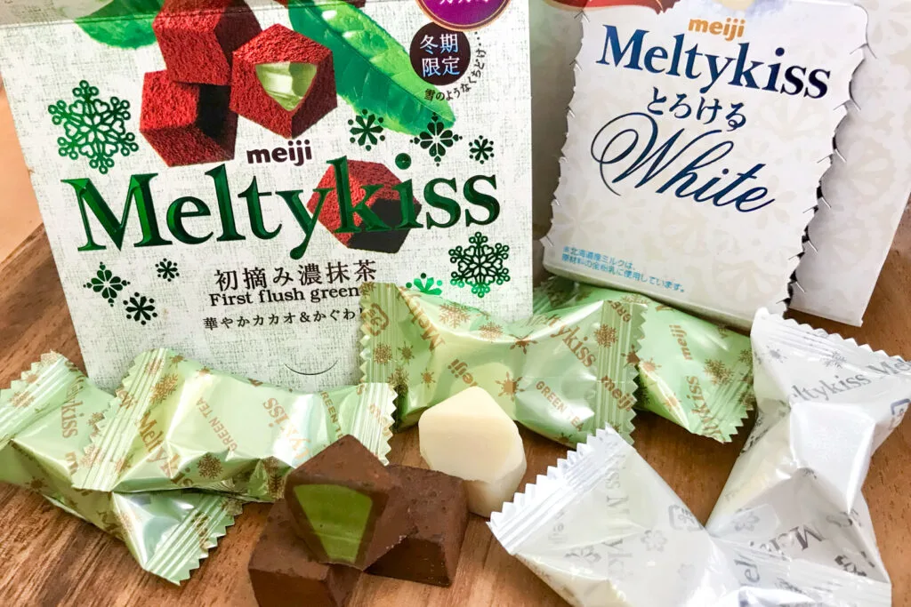 The cardboard packaging of Meltykiss in 'First flush green tea' flavor plus Meltykiss 'White'. In front of the packaging are a number of chocolates in their individual plastic wrappers (green tea in a light green color and the white in white packaging). There are also a few out of the packaging and one of each of them has a bite taken out it to show the inside. The green tea version has regular brown chocolate on the outside and a green center, and the white is white chocolate with a white center.