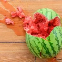 The aftermath of suikawari: A whole watermelon sitting on a wooden surface. The watermelon has been cracked open in a haphazard manner. The pink flesh can be seen between the top and bottom halves and some of the flesh and juice is spilling out towards the top left of the image.