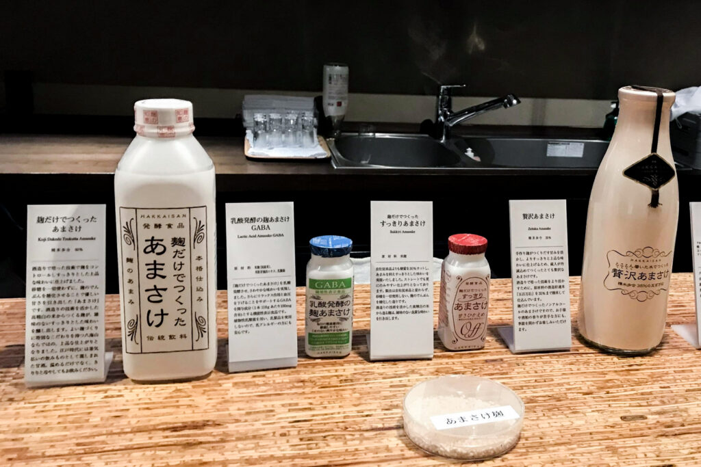 Four amazake (amasake) products lined up on a wooden bench at the Hakkaisan Brewery Tasting Room in Niigata. White labels with the product name and description are next to each of them. The room is dimly lit.
