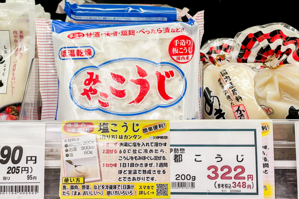 A Japanese supermarket shelf showing Iseso's Miyako Koji product. It is a clear sachet packaging that shows the white rice koji inside. The text on it is red with some blue design elements. The price is listed as 322 yen before tax and 348 yen inclusive of tax. On the product/price label is also an easy recipe for Shio Koji (salt koji).