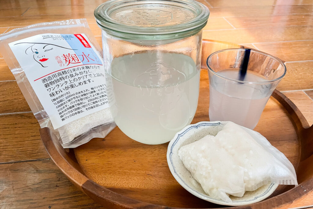 A sachet of koji rice packaged as a beauty product. Next to it is a glass jar with a glass lid that is about 2/3 full of the beauty tonic (koji rice plus water) and next to that is a small glass cup about 3/4 full with the tonic. In front of that is some more koji rice on a small white plate. It's all sitting on a round wooden tray on a wooden table.