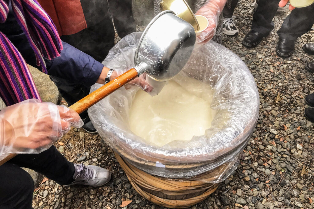 A plastic lined barrel of amazake with two people either side using large ladles that almost look like cooking pots with long handles to pour some into cups for visitors to a winter event.