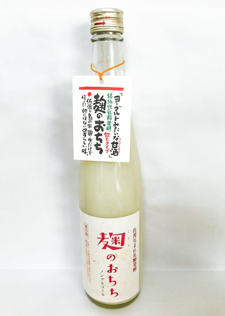 A bottle of Koji no Ochichi from Sado Island in Niigata Prefecture. The bottle is clear with a metal cap and has a white label with red writing. It is set on a white surface in front of a white background.