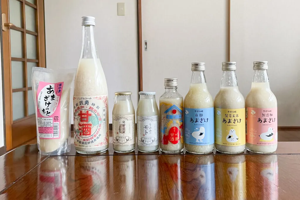 Eight types of Koji Amazake made by local sake breweries all lined up in a row on a wooden table in a Japanese-style room. The first one is a pouch and the others are varying size bottles. All the containers (pouch and bottles) are clear showing the light porridge-looking contents.