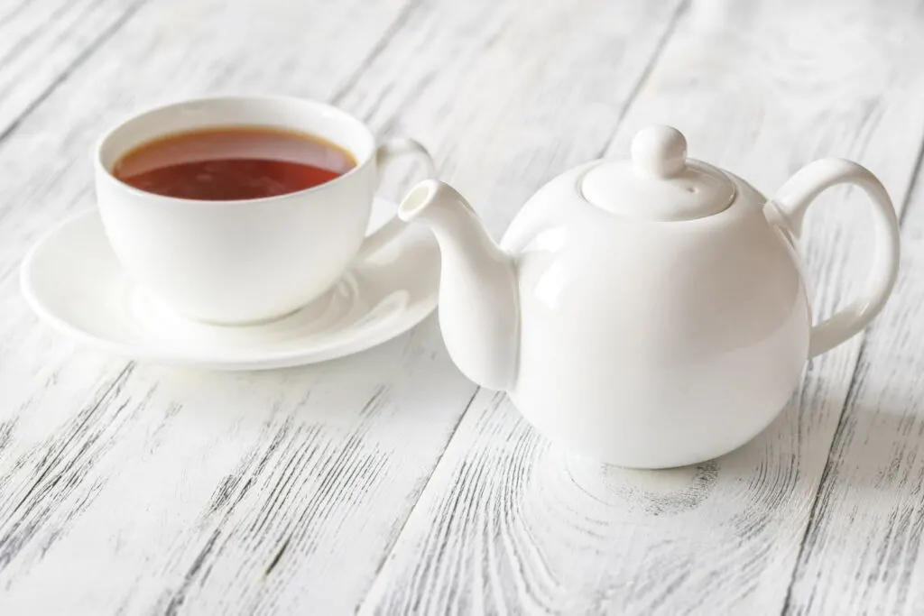 A white ceramic teapot to the right and a cup of "straight" black tea in a white tea cup on a white saucer to the left. They are placed on a wooden surface stained with a white paint.