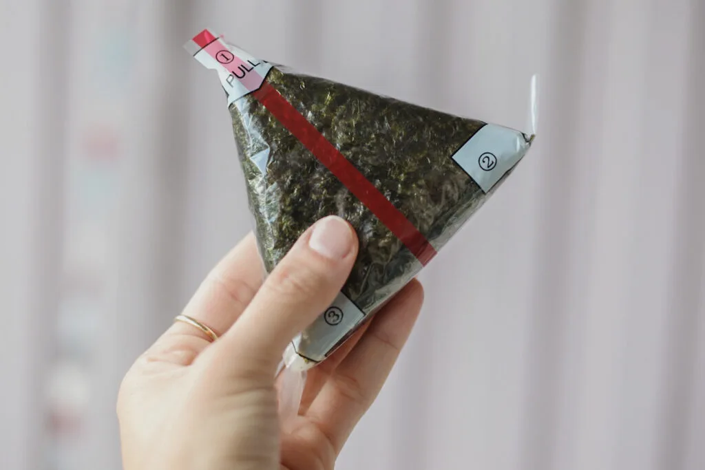 A left hand is holding up a triangle shaped onigiri wrapped in plastic from a convenience store. There is a red line down the center of the packaging and numbers 1-3 at each corner of the triangle to show the order of how to open it.