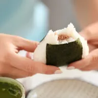 Japanese rice ball fillings: Manicured hands hold out a triangle-shaped Japanese rice ball with a rectangular piece of seaweed wrapped around it and a bite taken out of the top showing the tuna filling.