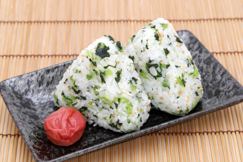 Two mazekomi onigiri (onigiri with an ingredient mixed throughout the rice). The onigiri filling is "aona" or a green vegetable. They are sitting on a speckled grey ceramic plate with one umeboshi (pickled plum) on the left. The plate is sitting on a bamboo mat.