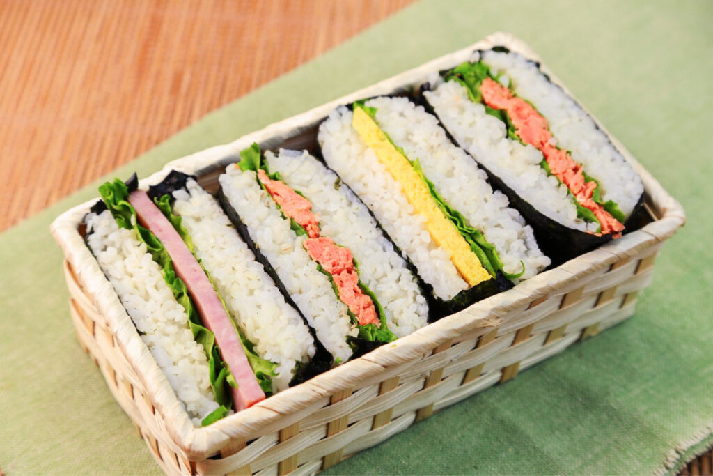 A small rectangular bamboo basket with four onigirazu inside that look like a sandwich cut in half, but instead of bread you have rice with seaweed on the outside. All the onigiri fillings have lettuce with one other ingredient. The first is Spam, the second is salmon, the third is egg and the last is also salmon. They sit on a light green table runner on a wooden table. 