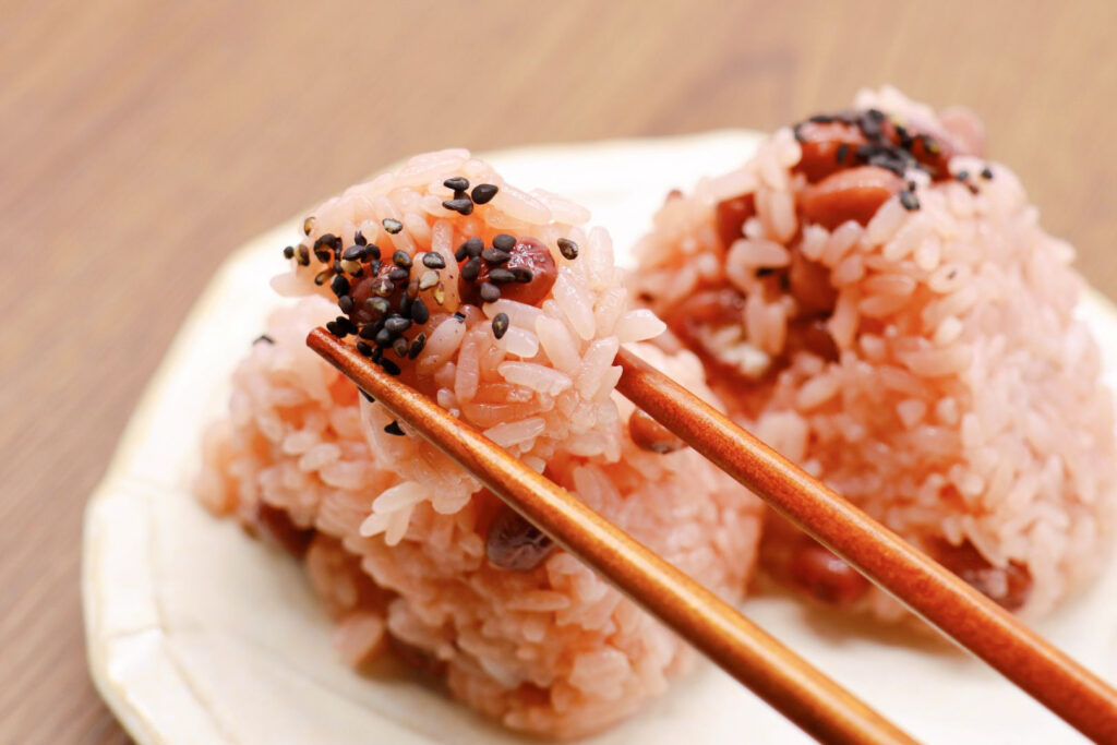 Two sekihan onigiri on a white ceramic plate. The onigiri rice is slightly red in color thanks to the red azuki beans and the onigiri are topped with a sprinkling of roasted black sesame seeds. A pair of chopsticks can be seen coming in from the right of the image and are holding up a mouthful of rice taken from the top of the onigiri on the left.