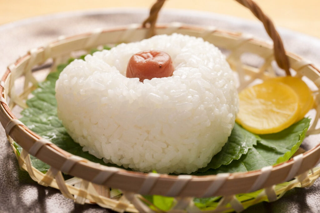 A large white rice triangle onigiri sits flat in a bamboo basket atop some green leaves. An umeboshi (pickled plum) has been pressed into the center of the onigiri from the outside and remains fully visible. Next to the onigiri are some pickles. 