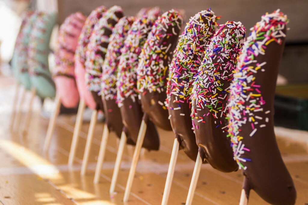 A row of choco-bananas on wooden sticks at a Japanese summer festival stall. The ones closest to camera are regular milk chocolate with multi-colored sprinkles. Pink and blue versions of the same treat can be seen further down the row.