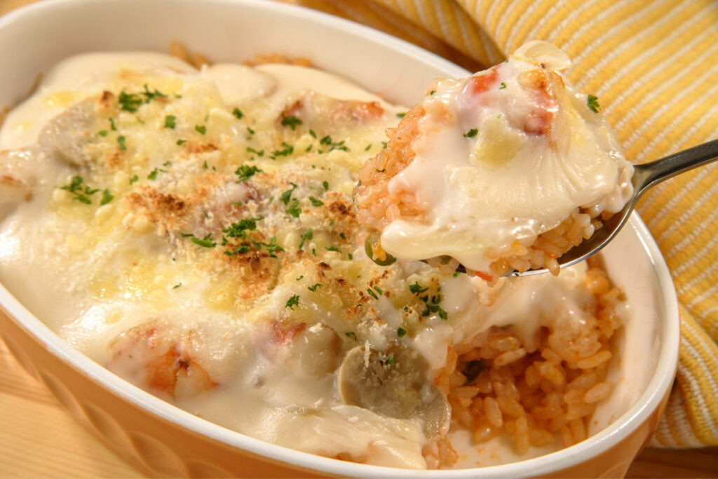 The first spoonful of a freshly-baked shrimp doria is being held up to the camera, revealing the rice underneath. Shrimp and mushroom are visible in the cream sauce, which is topped with cheese that has turned golden brown in the oven.