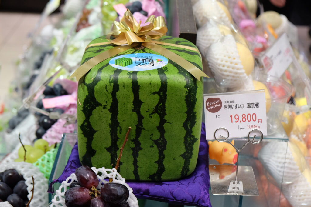 A square watermelon with a gold ribbon around it and the official Zentsujisan square watermelon sticker on top sits on a purple display cushion at the highest point in a high-end supermarket fruit display. The price next to it says 19,800 yen before tax (21,384 inclusive of tax).