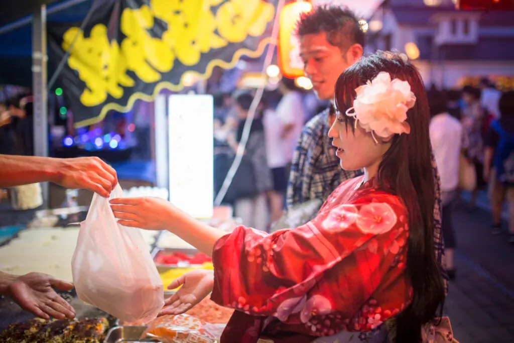 A young Japanese woman wearing a red yukata and a large pink flower accessory in her hair receives a plastic bag containing her okonomiyaki order from a stall vendor at a Japanese summer festival. It is already evening and her male companion can be seen next to her also wearing traditional Japanese summer festival garb.