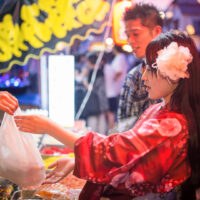 A young Japanese woman wearing a red yukata and a large pink flower accessory in her hair receives a plastic bag containing her okonomiyaki order from a stall vendor at a Japanese summer festival. It is already evening and her male companion can be seen next to her also wearing traditional Japanese summer festival garb.