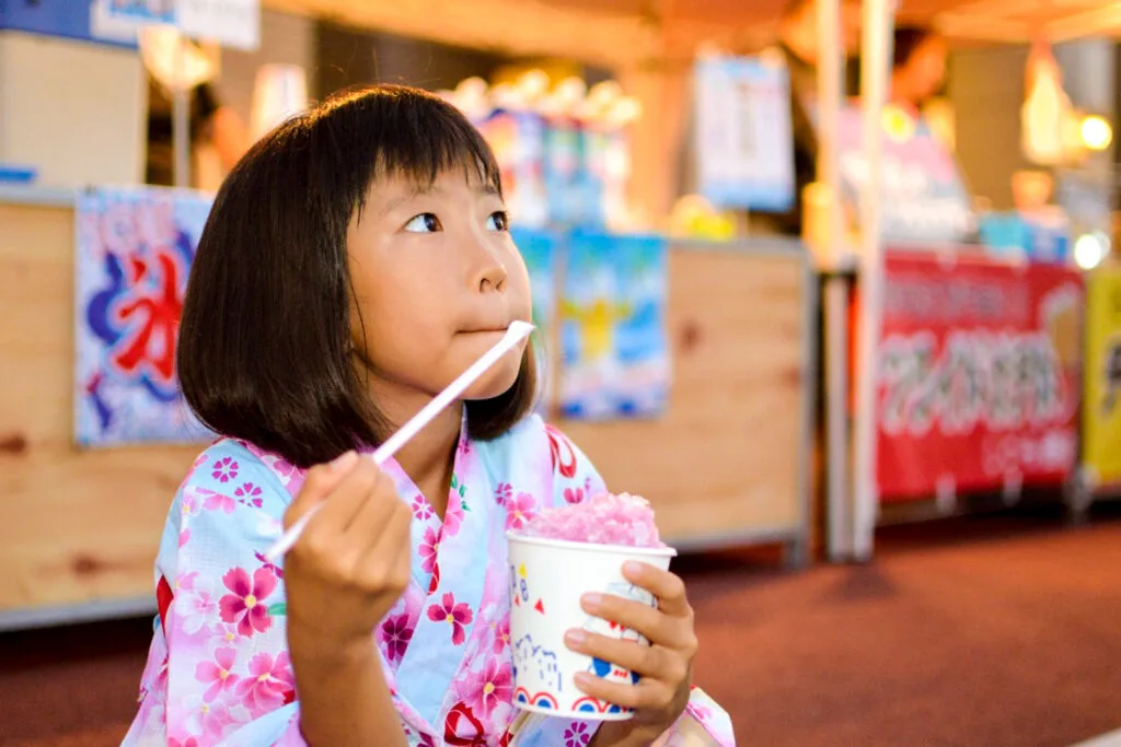 A young Japanese girl with a bob and fringe sits on a street kerb during a Japanese summer festival. She is wearing a jinbei and eating strawberry flavored kakigori with a straw with a scooped end. The kakigori stall can be seen in the background.