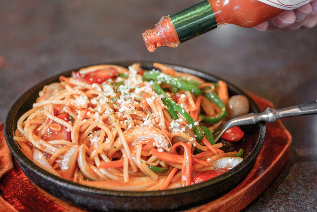 Tomato-based Napolitan spaghetti (Naporitan) being served in the saucepan it was cooked in. It already has a fork placed in it and has been sprinkled with some parmesan cheese. A hand can be seen adding some tabasco sauce.