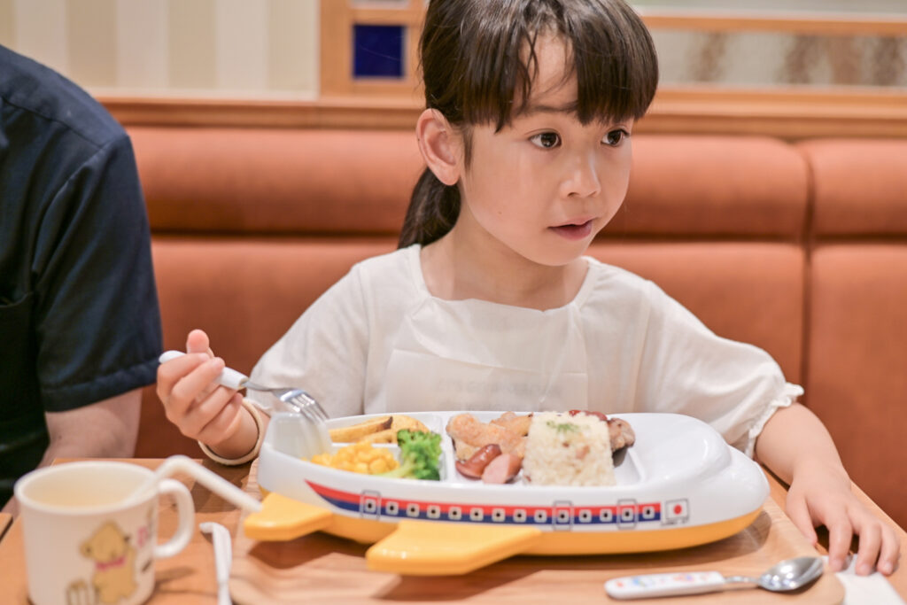 A young Japanese girl sits in a booth at a restaurant holding a fork in one hand with a kids meal in front of her. The plate is in the shape of an airplane, but it is not flat, it's more like a plastic toy that has compartments built into it on top for the food to go. Visible are fried wedges of potato, corn, broccoli, sausage and rice molded into the shape of Mt Fuji. We are unable to clearly see more of the main dishes behind the rice, but perhaps a hamburger patty and some fried chicken.