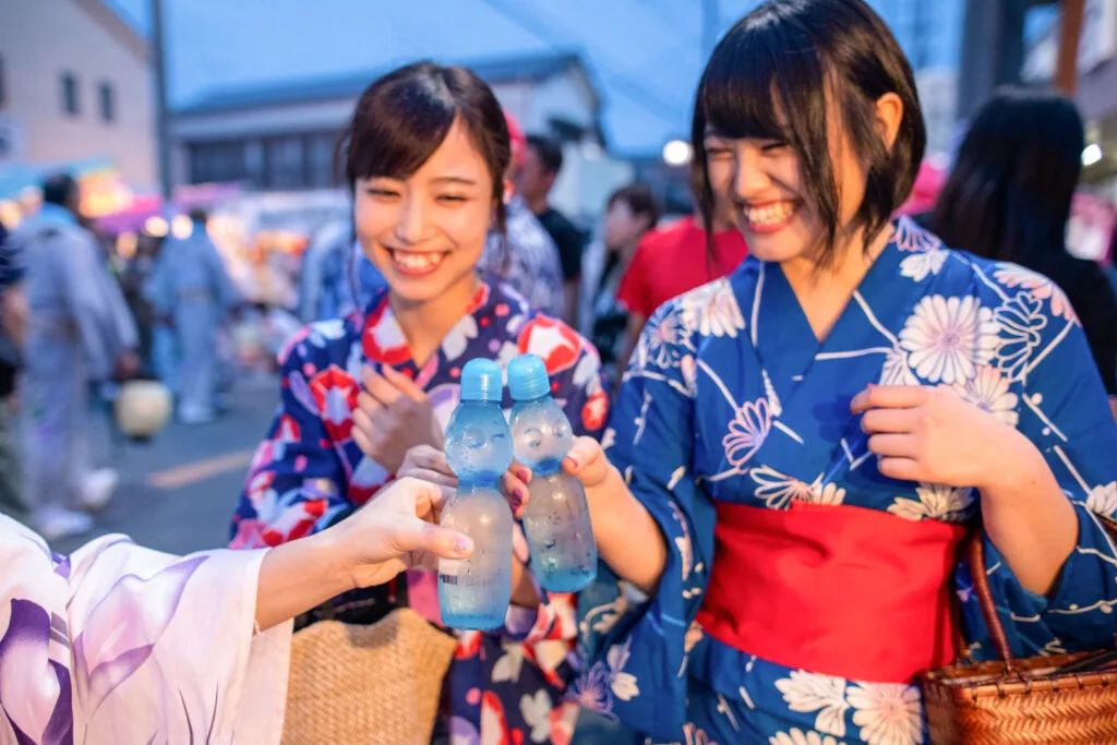 Two young Japanese women wearing yukata clink two bottles of ramune together in a 'cheers' motion at a Japanese summer festival. One of their faces is out of frame, while the other smiles widely. Another woman in the group watches on, also smiling.