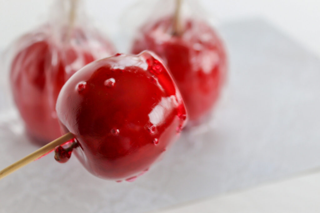 Three red candy apples on wooden sticks. Two are wrapped in clear plastic and sitting on a flat white surface in the background. The other one is unwrapped and being held up towards the camera from the left of frame.
