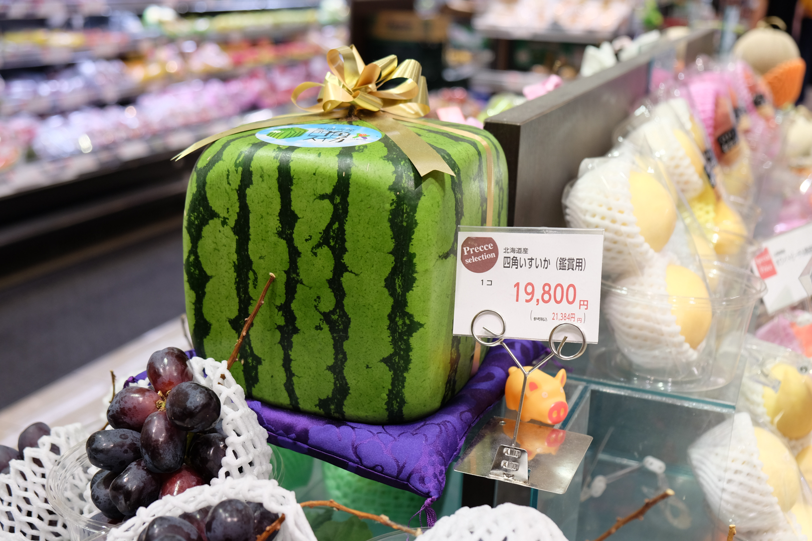 A square watermelon with a gold ribbon around it and the official Zentsujisan square watermelon sticker on top sits on a purple display cushion at the highest point in a high-end supermarket fruit display. The price next to it says 19,800 yen before tax (21,384 inclusive of tax).