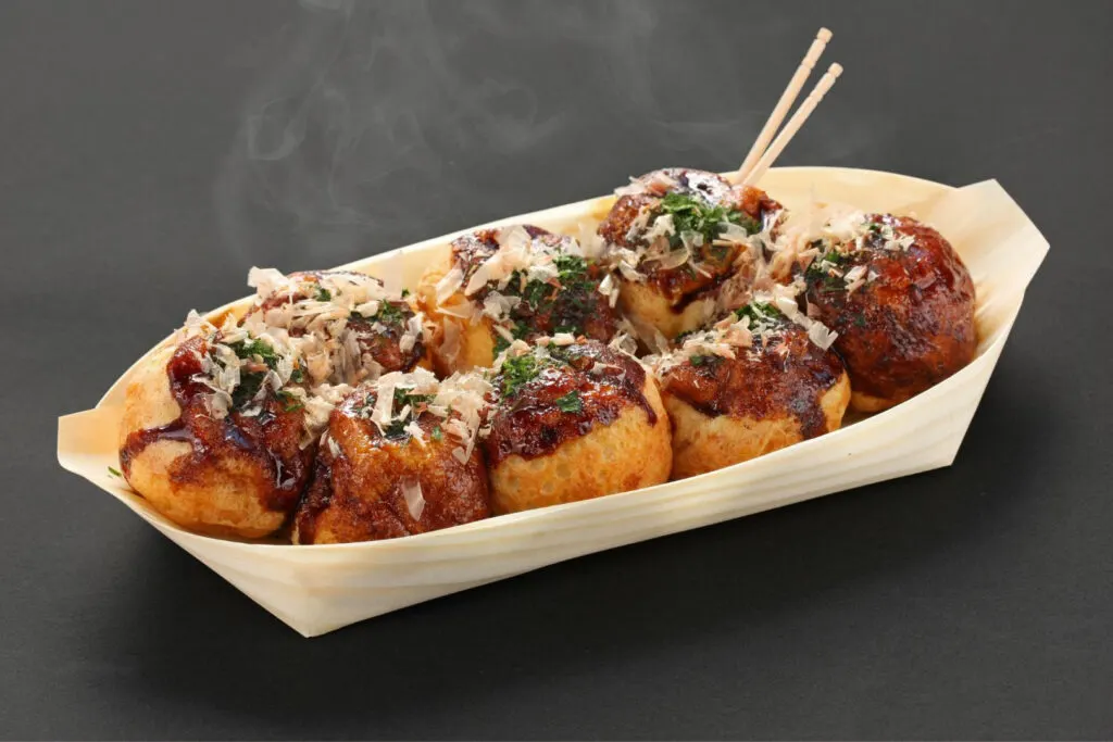 A paper take-out container with eight takoyaki covered in okonomiyaki sauce, aonori seaweed and bonito flakes sits on a black surface. Two toothpicks to eat them with are poked into one of the takoyaki (back-right).