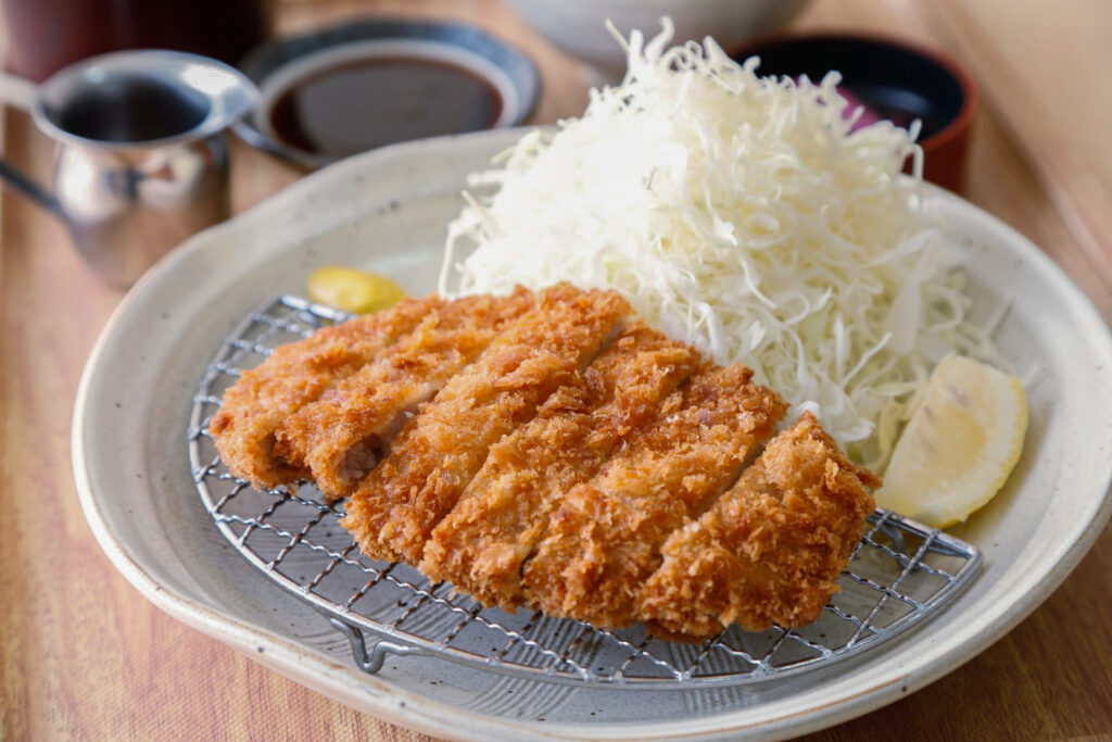 A sliced deep-fried pork cutlet on a dedicated half-moon shaped metal grid rack that fills half the plate, allowing air to circulate and therefore keeping the cutlet crispy throughout the meal. On the other half of the plate is a pile of cabbage, a wedge of lemon and a dollop of mustard. In the background, various other bowls and plates can be seen, including a small shallow dish of tonkatsu sauce.
