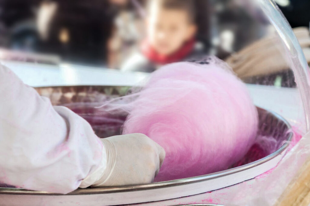 A photo of a wata-ame (cotton candy) vendor at a Japanese festival. The photo has been taken from the point of view of the vendor. You can see an arm with a white long-sleeved protective garment and white gloves preparing pink cotton candy on a stick. In the background, a child can be seen waiting amongst the crowd for some.