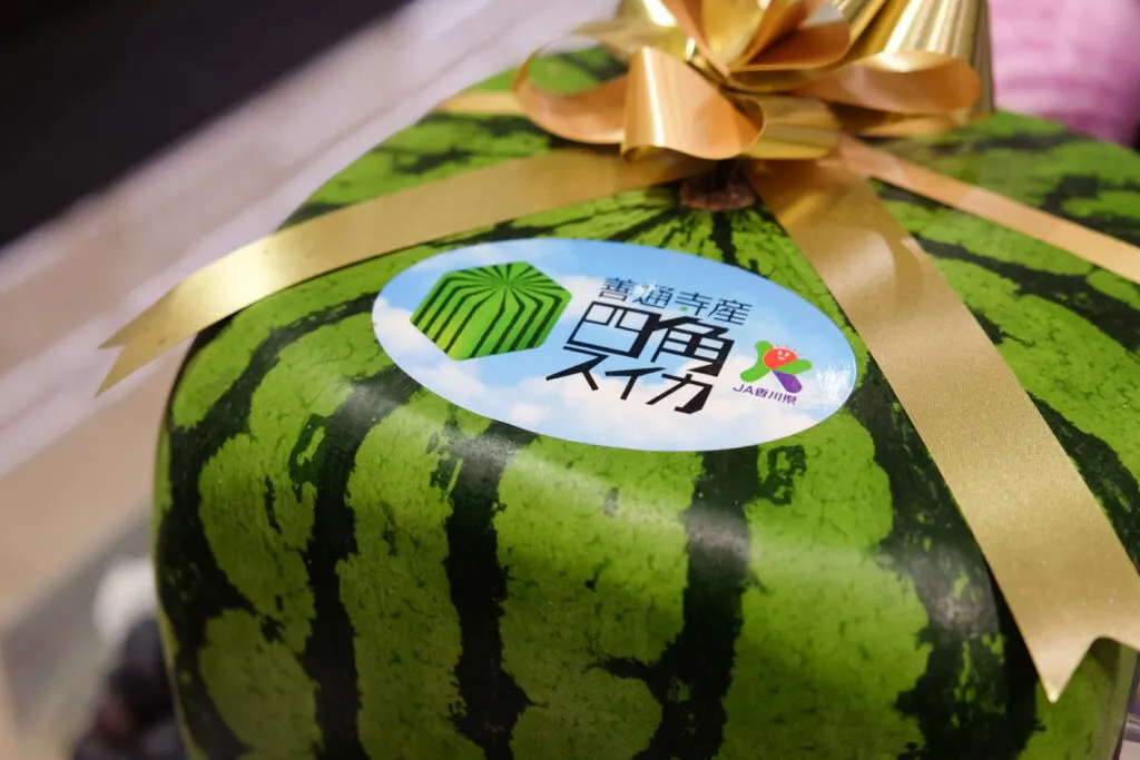 A close-up of the official Zentsuji square watermelon sticker. The background pictures a light blue sky with white clouds. To the left is a digital image of a square watermelon, then in the center in black text it says the name of the product 'Zentsujisan Shikakusuika' in Japanese, and to the right is the logo for Japan Agriculture (JA) Kagawa Prefecture, where Zentsuji is located.