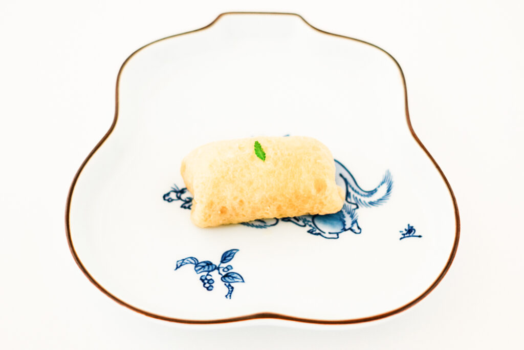One piece of dashi inari sushi from Kaiboku restaurant in Nihonbashi, Tokyo, with a small green leaf on top. It sits on a curvy white plate with a dark brown trim and an image of a fox in the middle in delft blue ink.