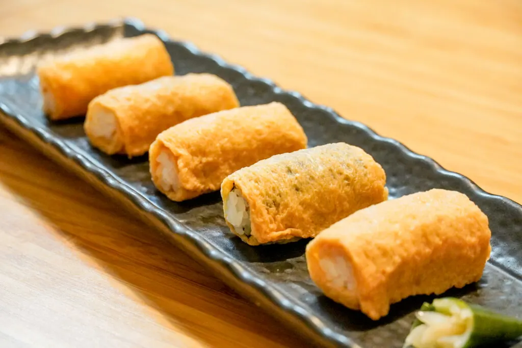 Five pieces of rolled up inari sushi (open at either end) using Kumamoto’s famous aburaage (Nankan-age). The pieces sit on a long black rectangular ceramic plate. Some pickles can also be seen on the plate closest to camera. It all sits on a plain light natural-colored wooden surface.