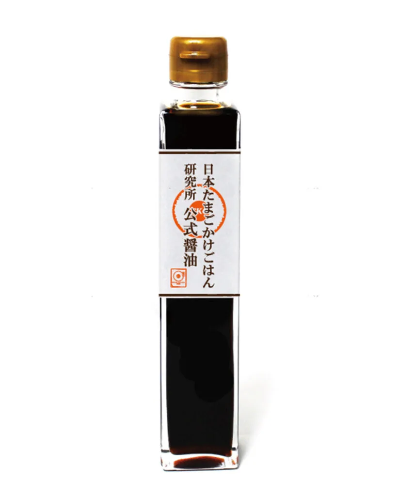 A tall and skinny bottle of the TKG Research Institute's official Tamago Kake Gohan soy sauce. The bottle is clear revealing the dark soy sauce inside with a white label wrapped around the middle - it has dark text and an orange logo on it. The plastic cap is gold in color.
