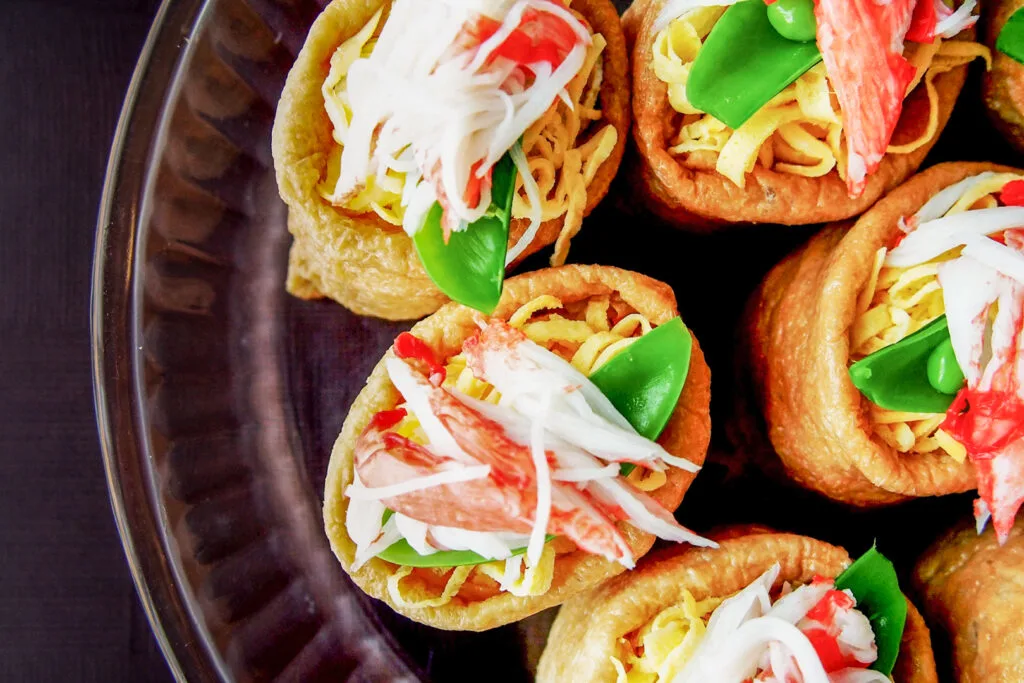 Inari sushi rolls with an open top (like a short burrito standing on end) on a dark brown ceramic plate with a decorative rim. Tucked in the opening is thinly sliced strips of egg, snap peas and shredded crab.