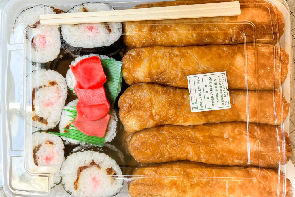 Five pieces of long inari sushi (shōdenzushi) and seven pieces of cut makizushi (sushi rolls) in a plastic takeout container with some bright pink-red pickled ginger and plastic bento grass.