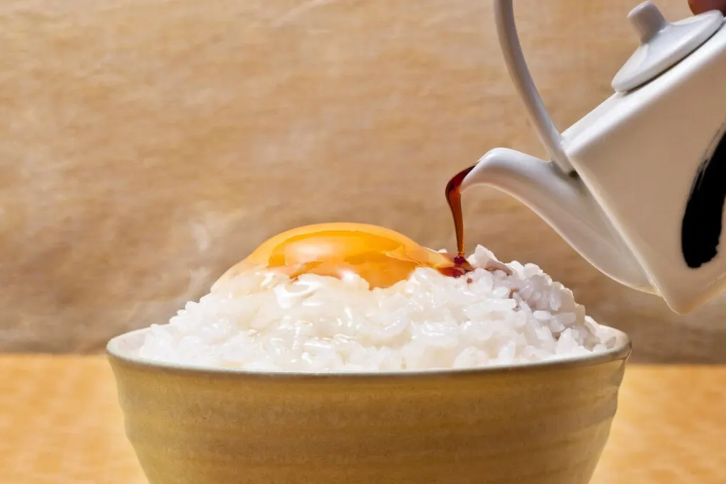 A white ceramic teapot with a blue design on it is pouring soy sauce onto a ceramic bowl of Tamago Kake Gohan.