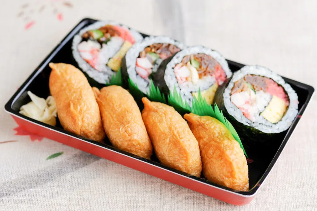 A 'sukeroku' pack with the lid off from a convenience store containing 4 inarizushi and 4 pieces of cut makizushi rolls. Some pickled ginger and plastic sushi grass (bento grass) can also be seen. It's sitting on a white table cloth with a subtle botanical design.