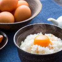 A dark ceramic bowl of Tamago Kake Gohan on a blue tablecloth. A perfectly intact raw egg yolk sits on top of the white rice. Soy sauce can't be seen in the dish, although there is a white ceramic soy sauce dispenser to the right. Behind the TKG is a large bowl of uncracked eggs and to the left we can see a small bowl with the egg shell from the egg that's already in the dish.
