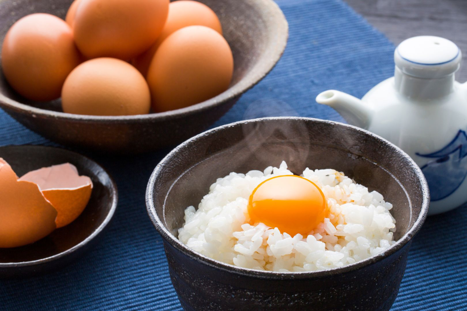 A dark ceramic bowl of Tamago Kake Gohan on a blue tablecloth. A perfectly intact raw egg yolk sits on top of the white rice. Soy sauce can't be seen in the dish, although there is a white ceramic soy sauce dispenser to the right. Behind the TKG is a large bowl of uncracked eggs and to the left we can see a small bowl with the egg shell from the egg that's already in the dish.