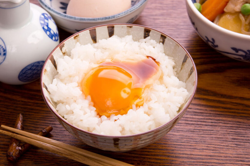 A bowl of Tamago Kake Gohan (TKG) in a ceramic bowl with organic brown stripes. The bowl is filled with white rice and has a raw egg on top with a little soy sauce. In front of the bowl are some wooden chopsticks on an organically-shaped wooden chopstick rest that looks like the fork of a tree branch, and around the bowl, various other dishes are partially visible in the shot, including a ceramic dispenser for soy sauce.
